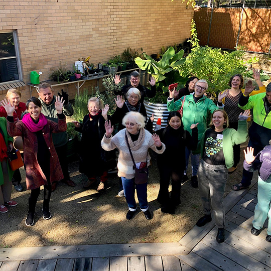 A dozen-strong group of people smiling and waving in the direction of the camera. They are in a small yard surrounded by plants and greenery.